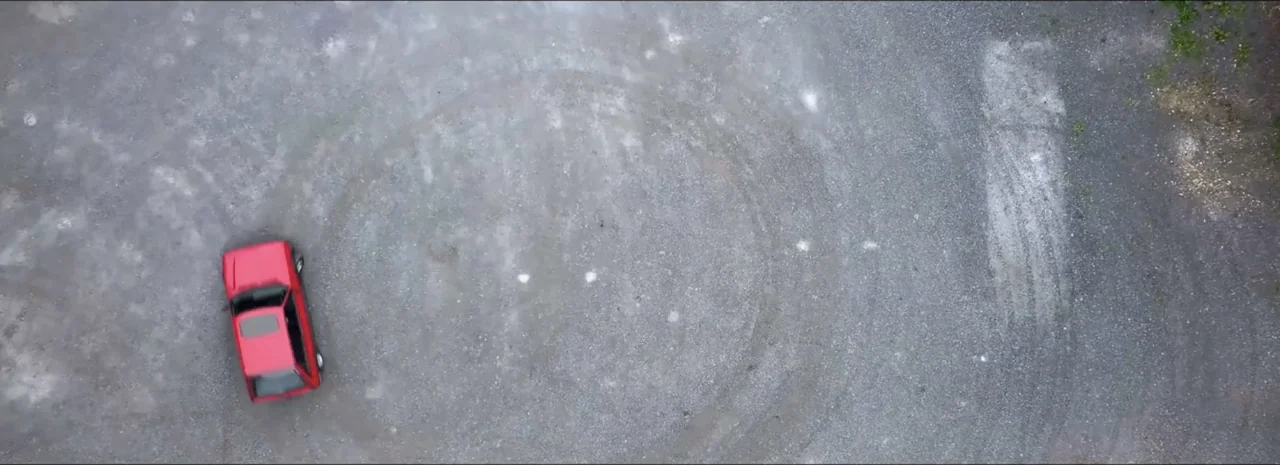 DannyBoy (2020) movie still: wide — A vast expanse of grey gravel is marked by circular tire tracks. A single red car sits parked in the lower left…