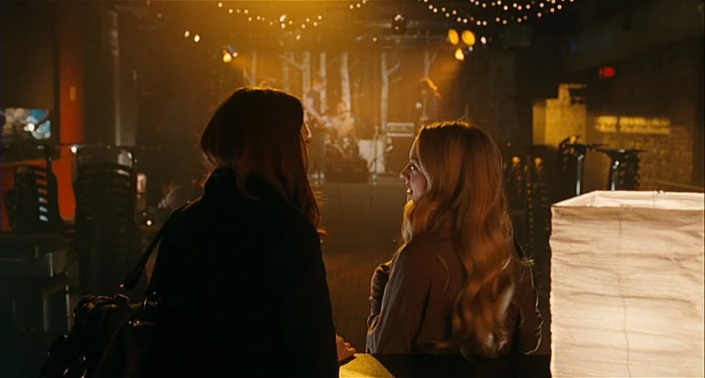 Chloe (2009) movie still: medium — A dark, dimly lit venue features a bar area with warm overhead lighting strings. Two women stand at the counter…