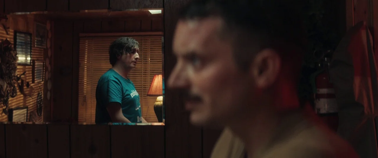 Come To Daddy (2019) movie still: medium — A dark, wood-paneled room is dimly lit by a lamp sitting on a surface. A man in a teal shirt is reflected…