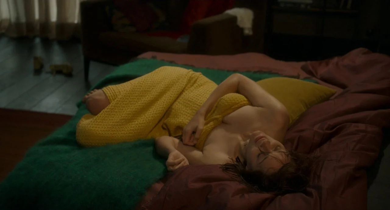 Annette (2021) movie still: medium — A dimly lit room with dark bedding and a green throw blanket draped over. A woman rests on the bed while…