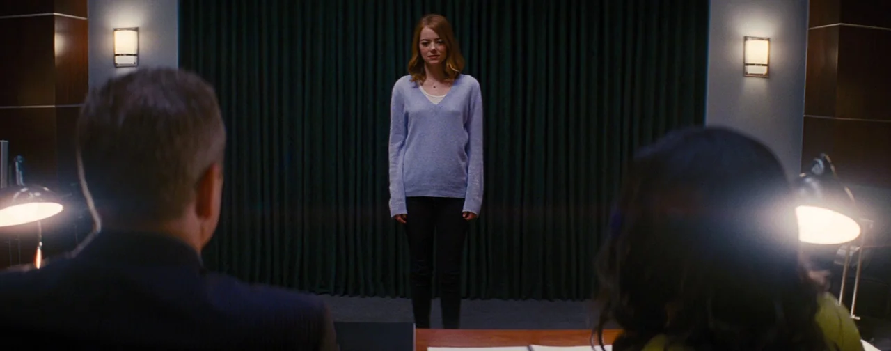 La La Land (2016) movie still: fullbody — A conference room with a dark green curtain background and two desk lamps. A young woman stands before two…