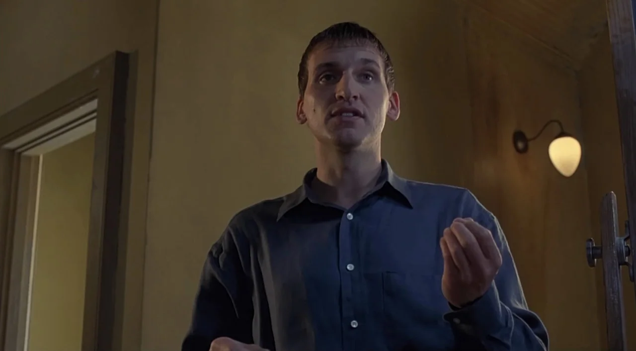 eXistenZ (1999) movie still: medium — An dimly lit interior hallway with plain walls features a wall lamp. A man stands in the center gesturing as…