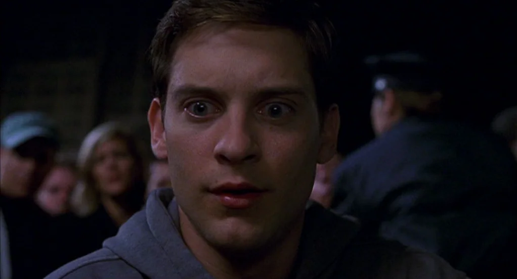 Spider-Man (2002) movie still: closeup — A dark, blurred outdoor space is filled with indistinct people standing around at night. A young man stares…