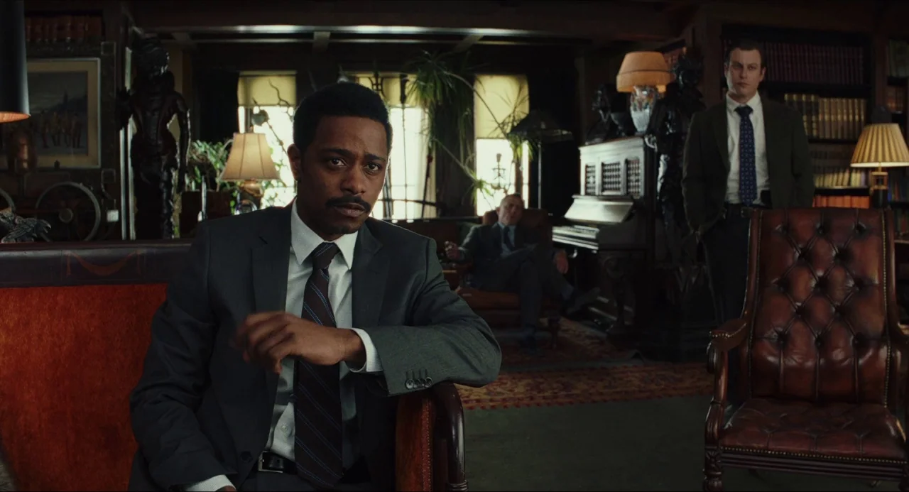 Knives Out (2019) movie still: medium — A dimly lit, wood-paneled room is filled with bookshelves and leather furniture. Three men in professional…