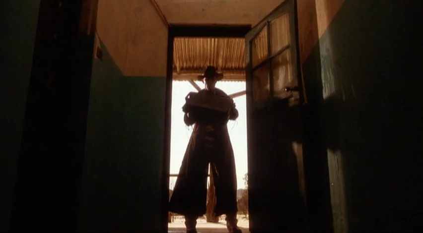 Dust Devil (1992) movie still: medium — A dimly lit narrow hallway with teal-painted walls leads to an exterior doorway. A man in a long coat and…