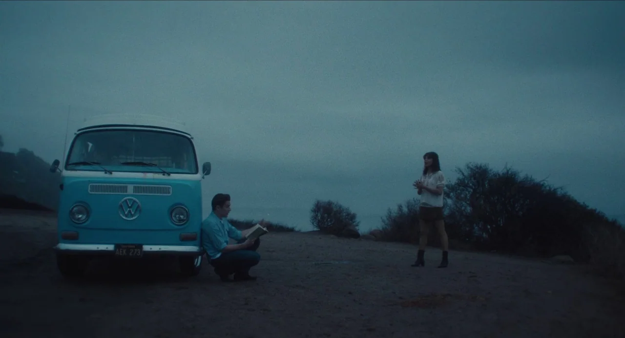 Born To Be Blue (2015) movie still: wide — A gravel turnout overlooks a dark ocean under a heavy, overcast twilight sky. A man crouches by a light…
