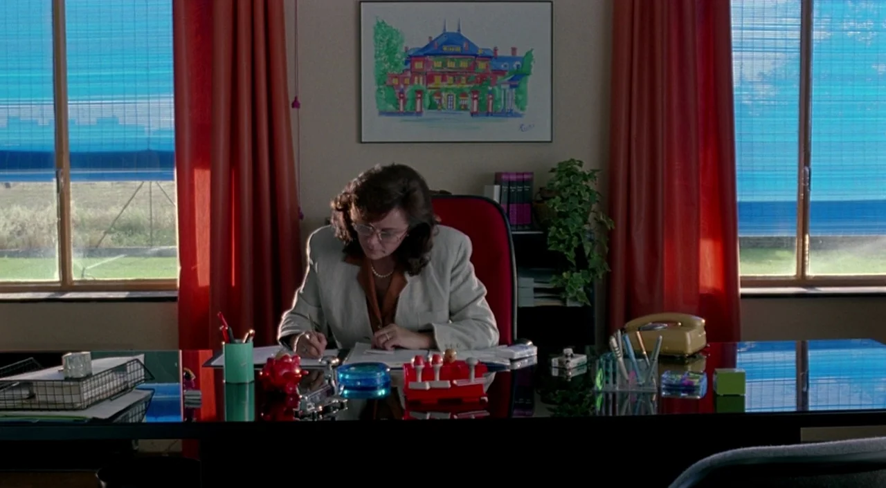 Tie Me Up! Tie Me Down! (1989) movie still: medium — A woman works at a glass-topped desk in a room with bright red curtains. Framed architectural…