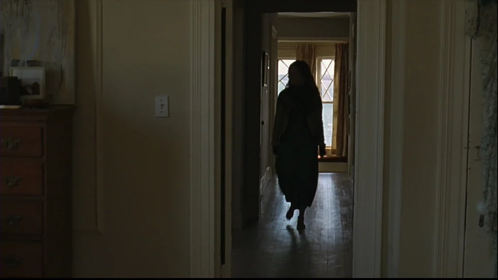 Margot At The Wedding (2007) movie still: medium — A dimly lit interior hallway with hardwood floors and beige walls. A woman walks toward the…