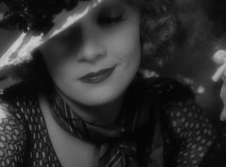 Blonde Venus (1932) movie still: closeup — A woman wears a vintage hat that casts a deep shadow across her eyes. She holds a cigarette as her eyes…