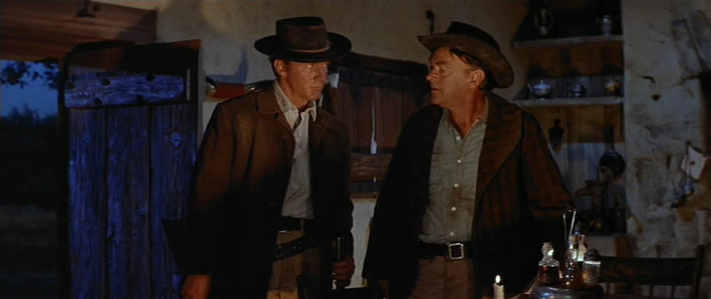 Flaming Star (1960) movie still: medium — A dimly lit rustic cabin interior has wooden walls and simple shelves with metalware. Two men wearing…