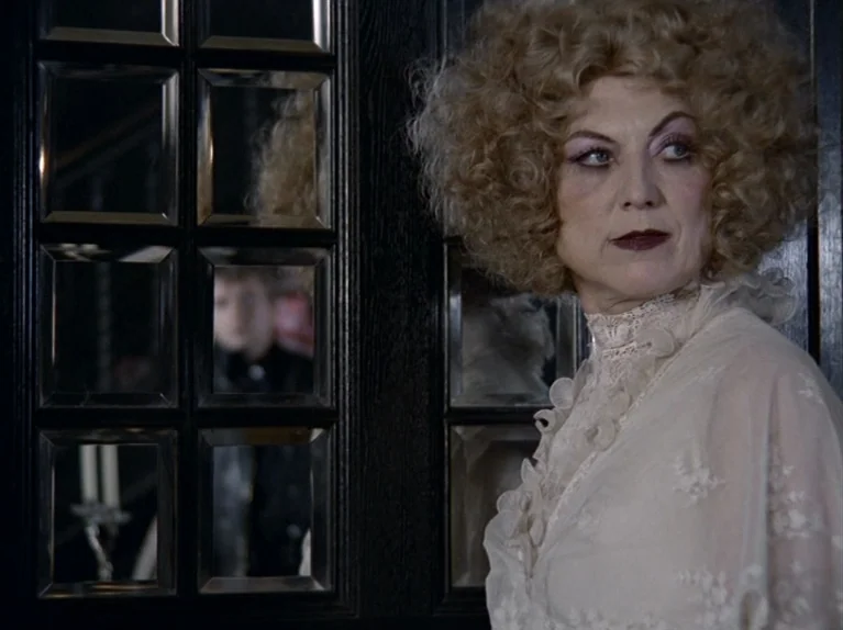 The Third Generation (1979) movie still: medium — A woman stands in a dark hallway featuring a black wall with mirrored panes. A blurry reflection…