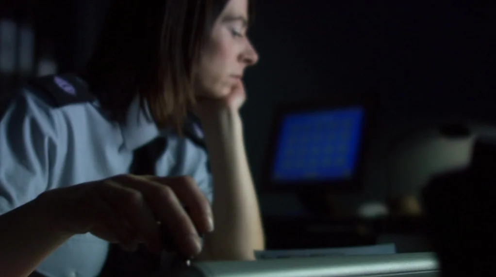 Red Road (2006) movie still: medium — A dimly lit security station contains a woman sitting at a dark workspace. The monitor screen in the…