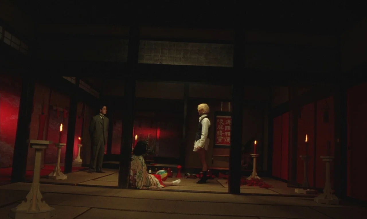 Yumeji (1991) movie still: wide — A dark, traditional Japanese interior features tatami mat floors and red paper walls. Three figures occupy the…