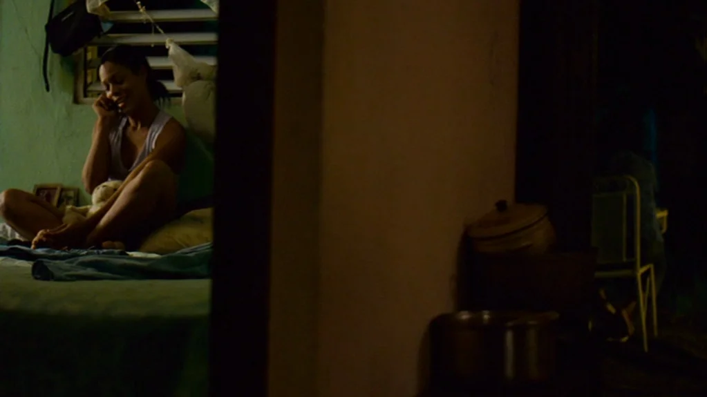 Sugar (2008) movie still: medium — A dimly lit room with green walls contains a bed where a woman sits. She holds a mobile phone to her ear while…
