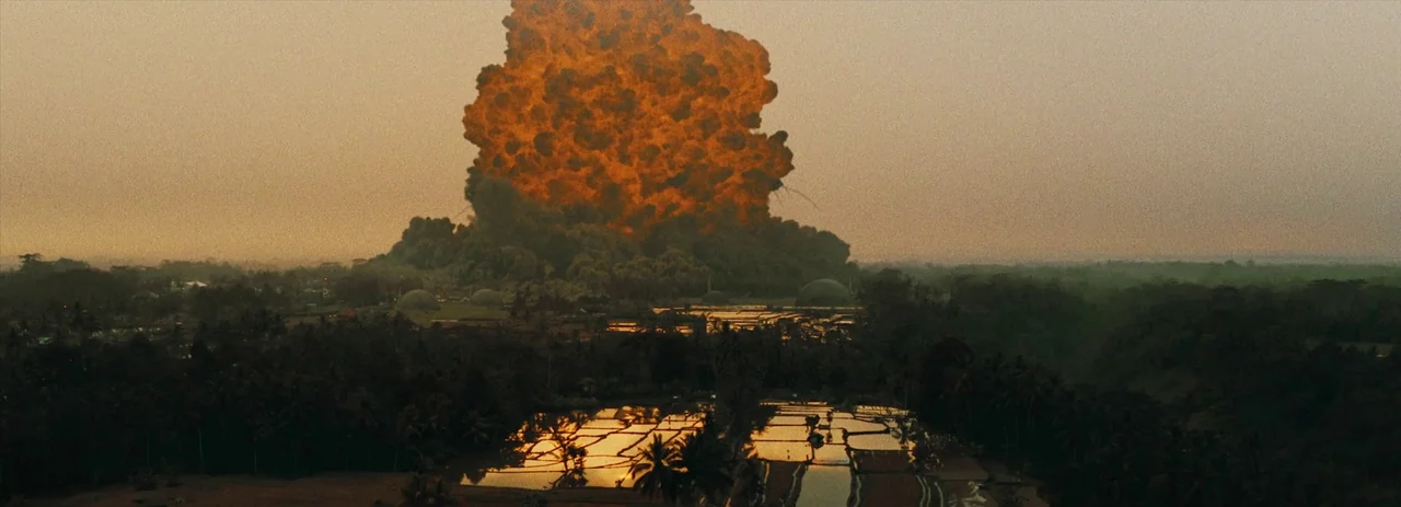 The Creator (2023) movie still: wide — A massive fire plume rises into the sky over a rural landscape of flooded rice paddies. The explosion…
