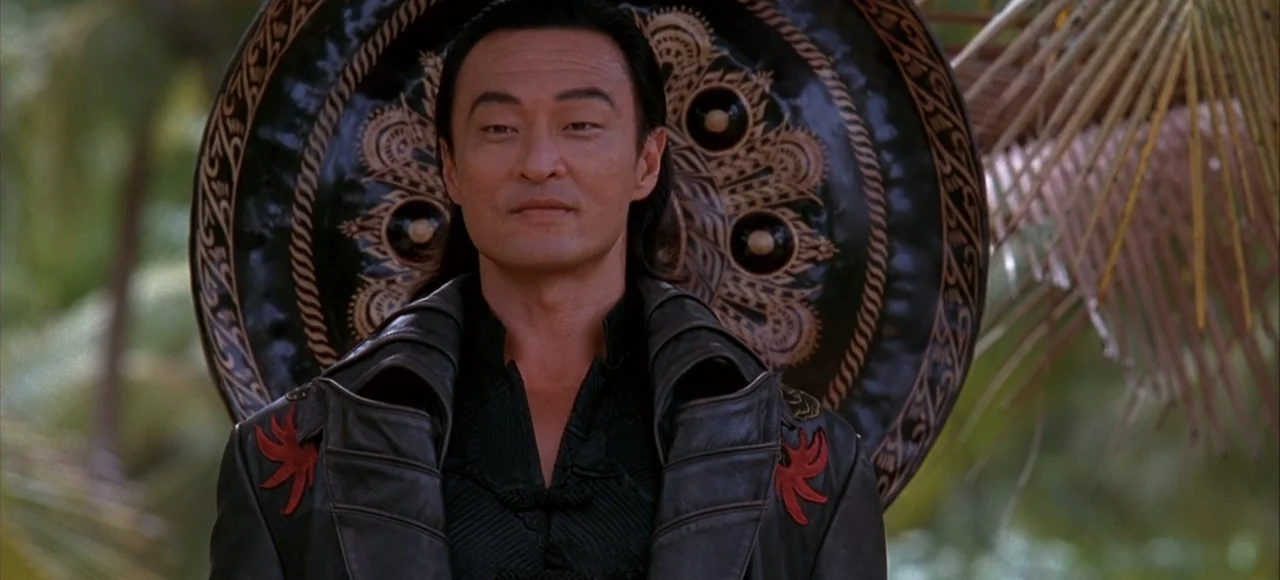 Mortal Kombat (2021) movie still: medium — A man stands outdoors in front of a large decorative dark circular gong. The background is blurred with…