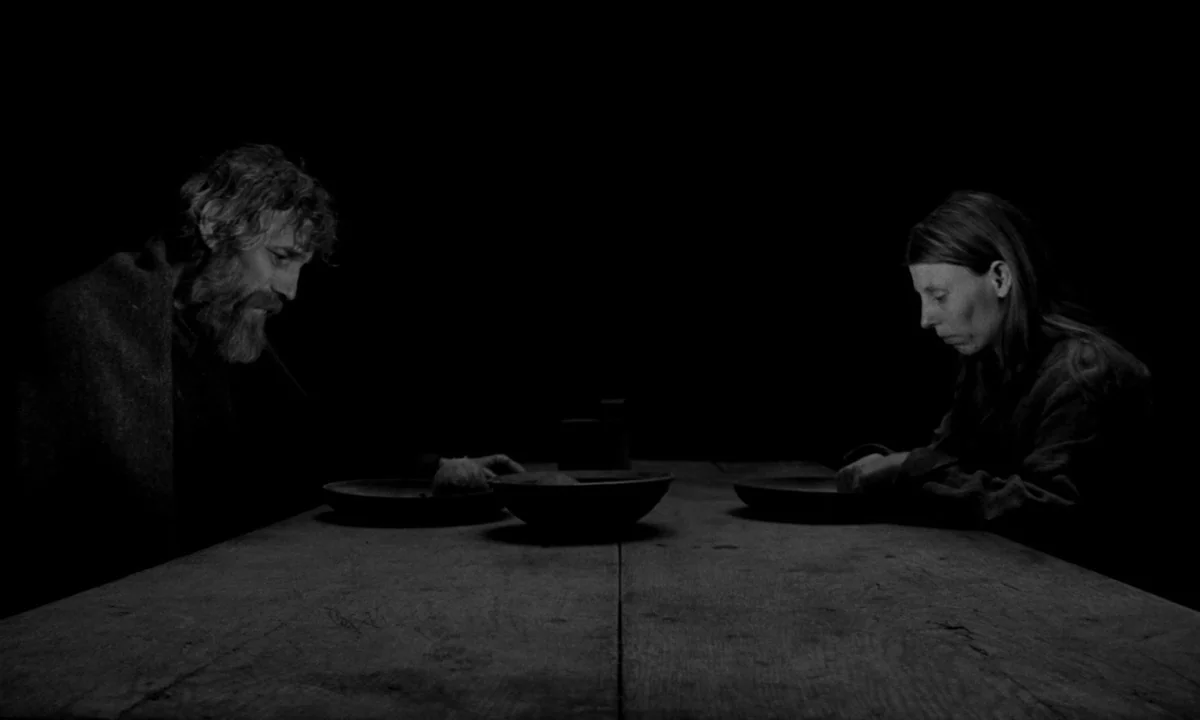 The Turin Horse (2011) movie still: medium — A dark, spartan interior with a long, plain wooden table centered in the frame. A man and a woman sit…