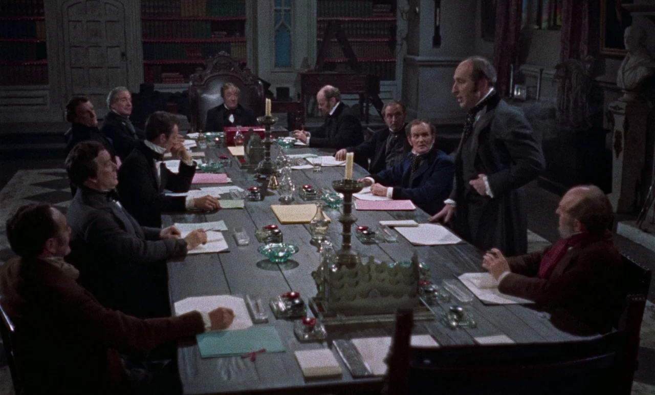 The Revenge of Frankenstein movie still: wide — A dimly lit room features a large, long wooden table surrounded by seated men. One man stands at the…
