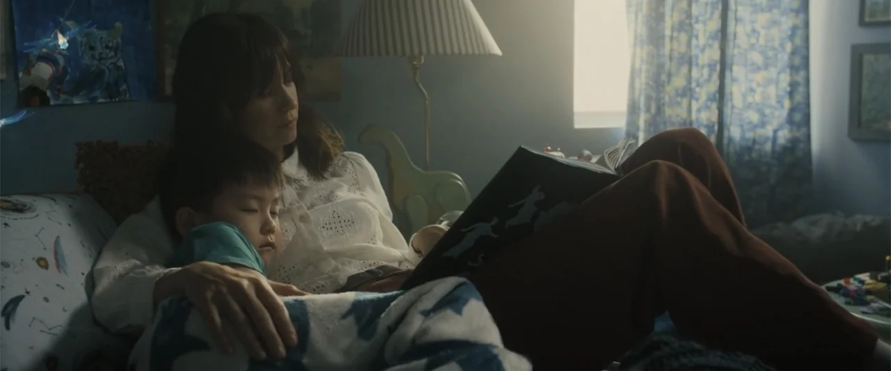 Afraid (2024) movie still: medium — A quiet bedroom contains a bed with a printed quilt and a bedside lamp. A woman sits on the edge of the bed…