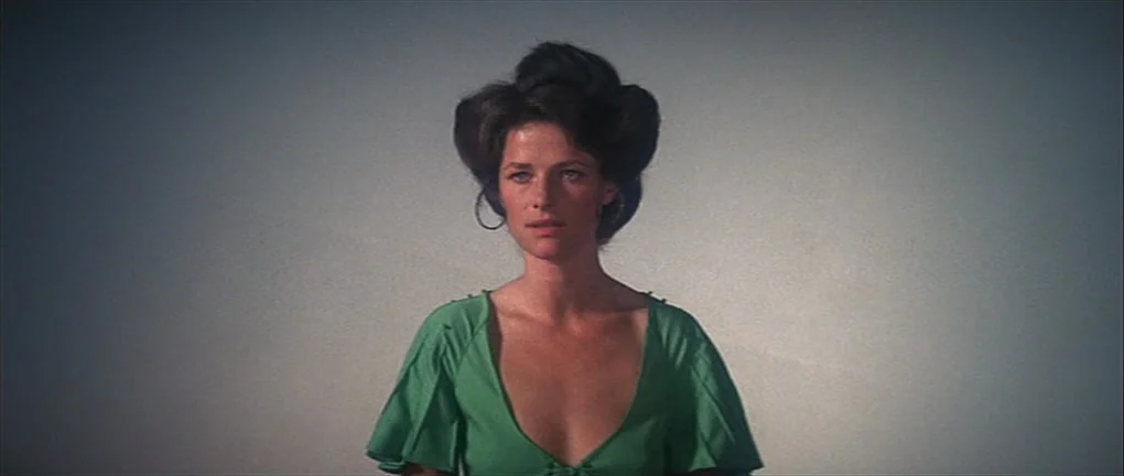 Zardoz (1974) movie still: medium — A woman stands in front of a featureless, light-colored studio wall. The woman gazes forward while wearing a…
