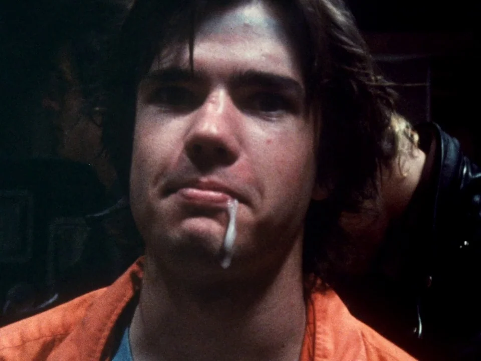 Decline of Western Civilization (1981) movie still: closeup — A dark indoor space featuring a man wearing a bright orange uniform. He stares ahead…