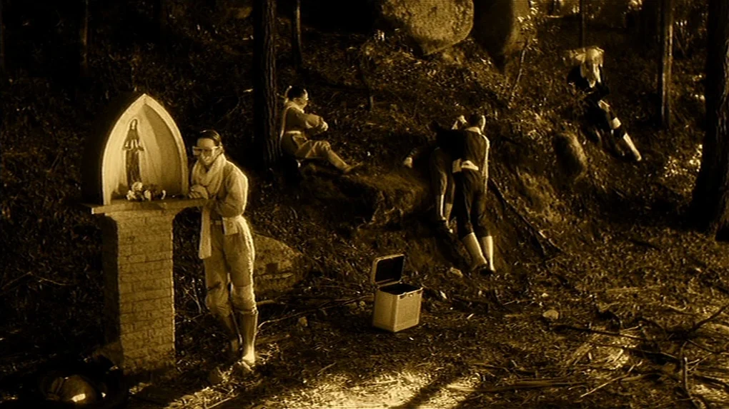 The State of Things (1982) movie still: wide — A group of people occupies a dirt hillside beneath tall pine trees. A stone shrine with a statue…