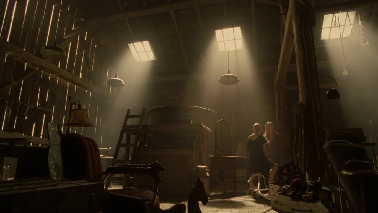 Splice (2009) movie still: wide — A cluttered barn interior is filled with vintage furniture, old trunks, and wooden beams. Two women stand in the…