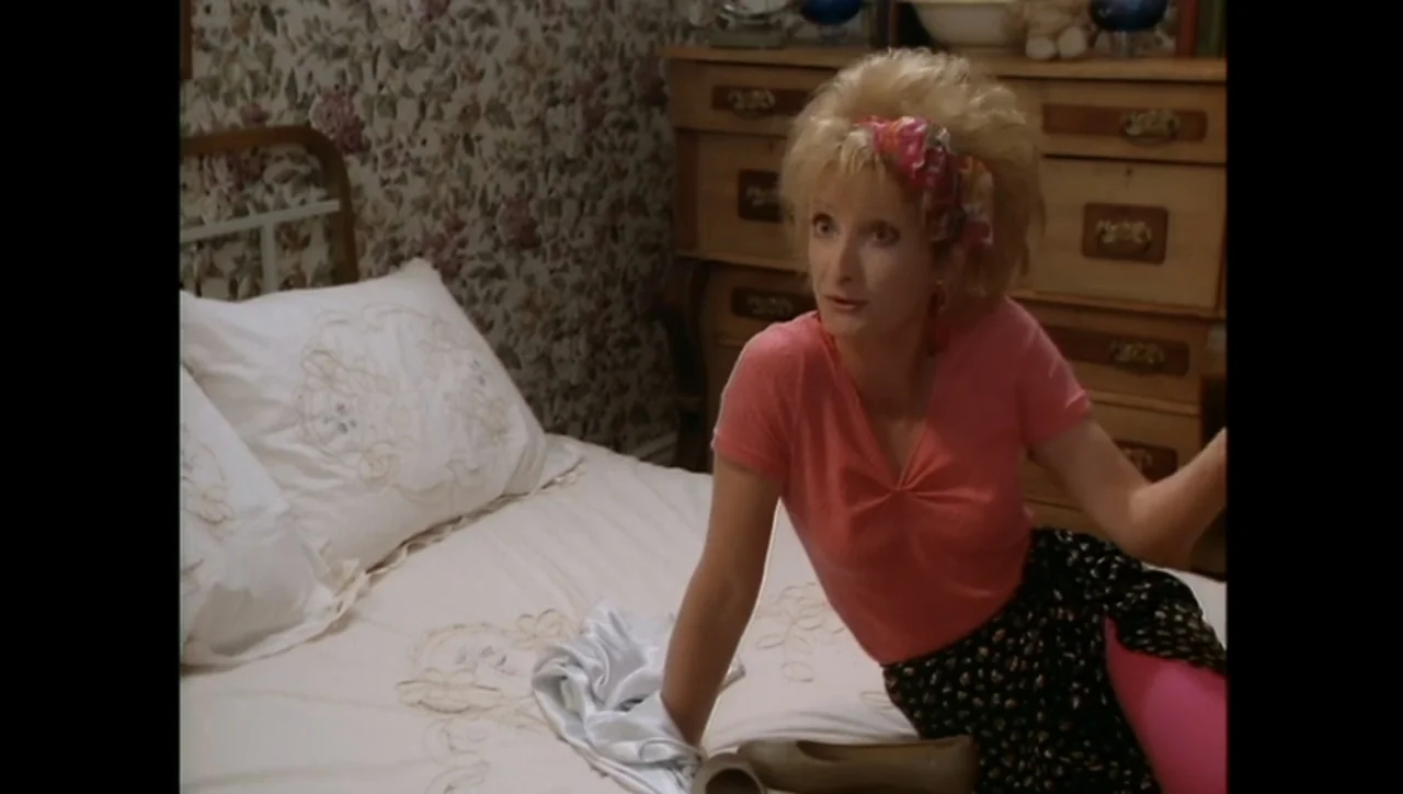 The Awakening (1995) movie still: medium — A 1980s bedroom features floral wallpaper, a white bed, and a wooden chest. A woman sits on the side of…
