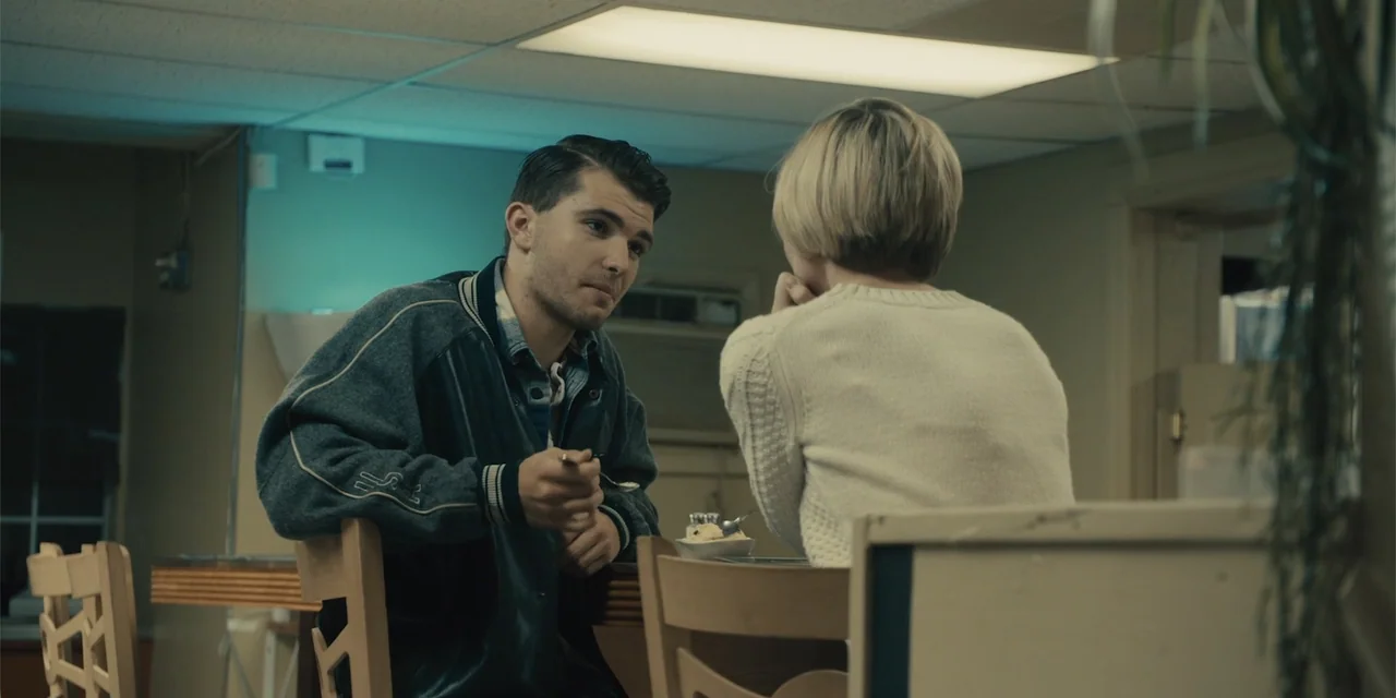 All Roads to Pearla (2019) movie still: medium — A quiet diner interior with dull beige walls and overhead rectangular fluorescent lighting panels.…