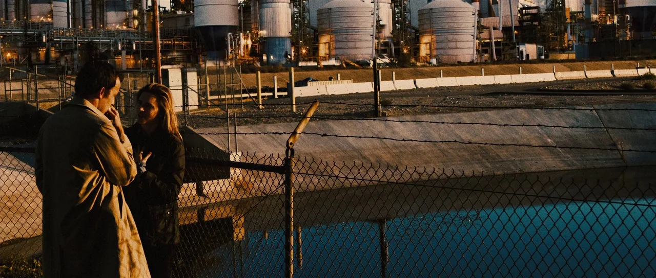 The East (2013) movie still: wide — An industrial facility with large storage tanks looms behind a barbed wire chain-link fence. A man and a woman…