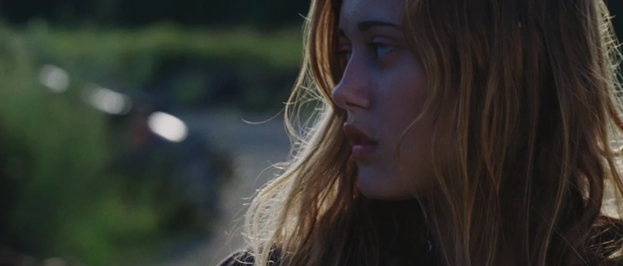 Wildlike (2014) movie still: closeup — A person stands in an outdoor area with blurred green vegetation in the background. The individual's face is…