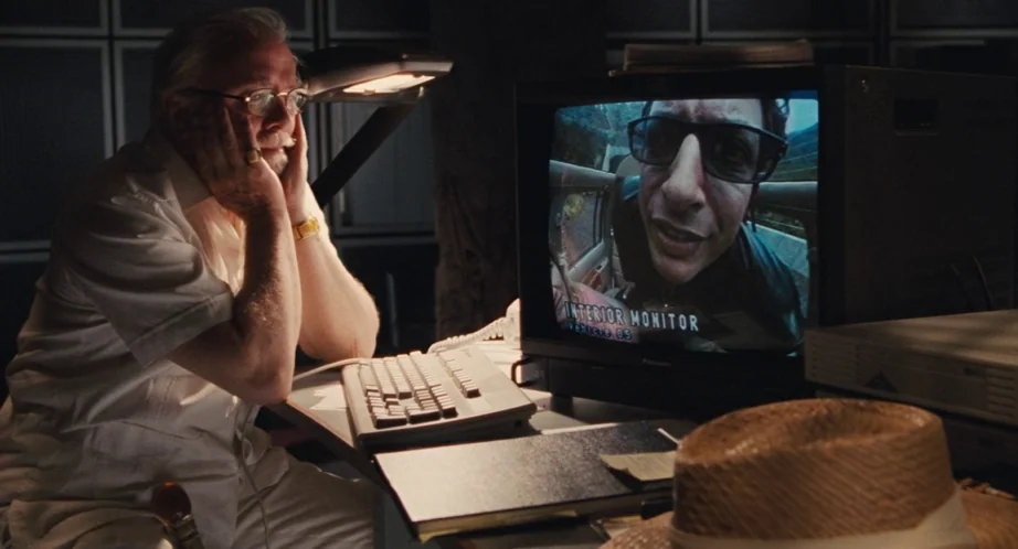Jurassic Park (1993) movie still: medium — A dark, dimly lit control room contains a computer monitor and various desk items. An elderly man in a…