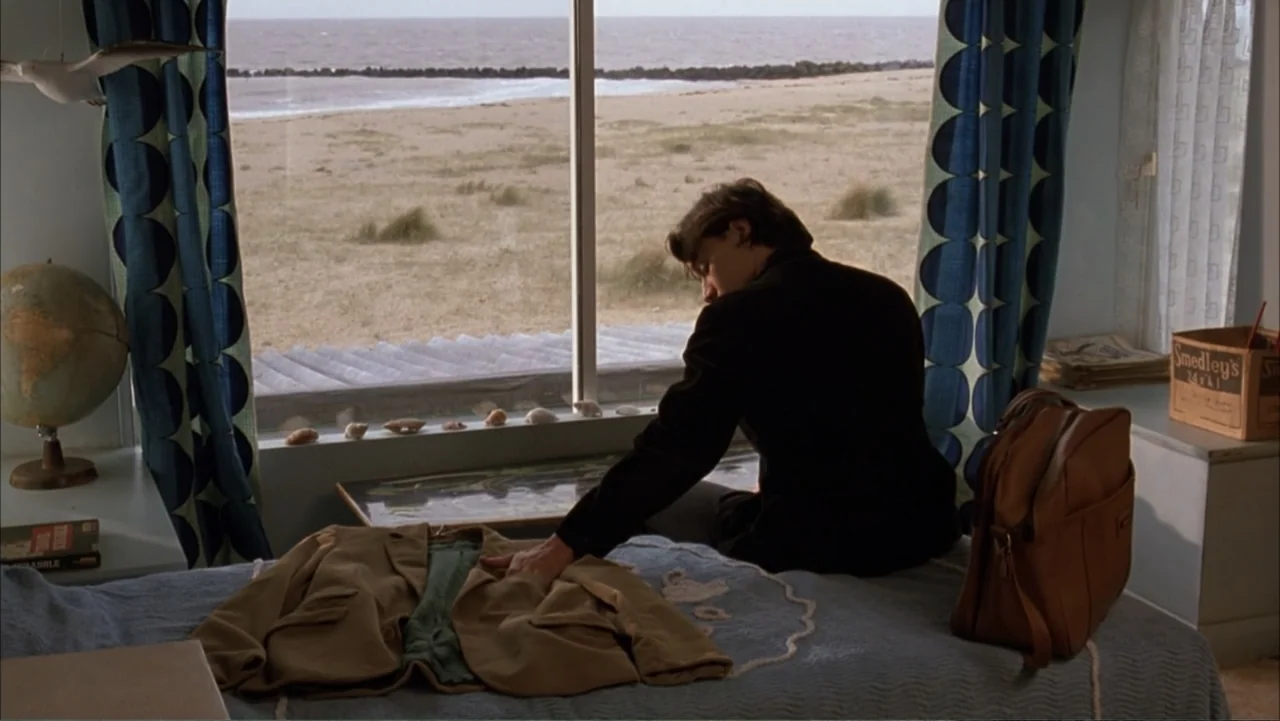 Starter for 10 (2006) movie still: medium — A seaside room features a large window overlooking a sandy beach and ocean. A man sits on a bed with a…