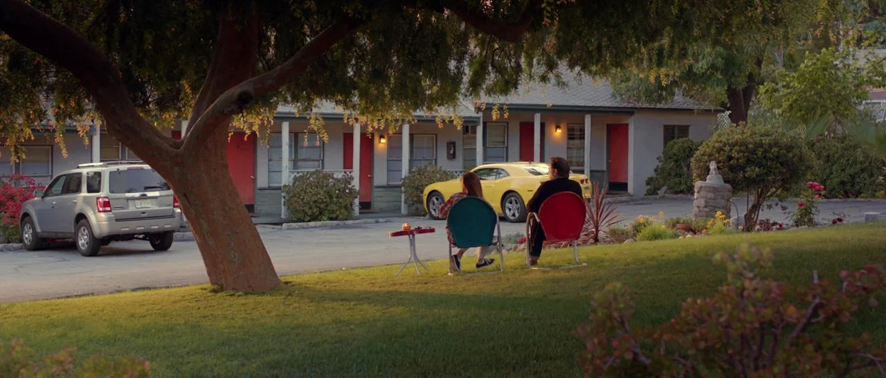God Bless America (2011) movie still: wide — A motel courtyard features a paved lot with a silver SUV and yellow car. Two people sit in colorful…
