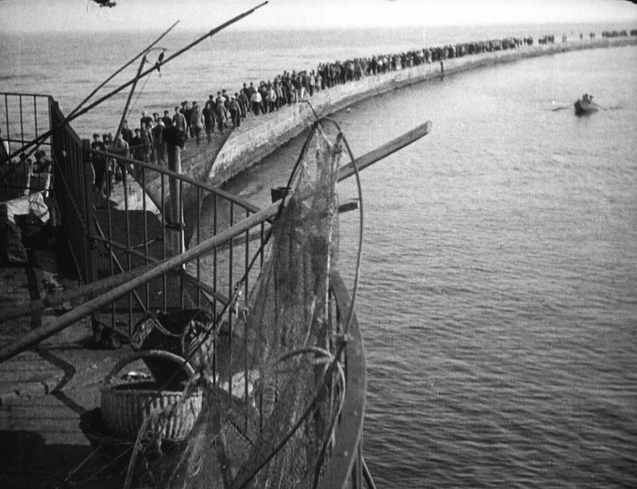 Battleship Potemkin (1925) movie still: wide — A long, curved concrete breakwater stretches into calm water under an overcast sky. A large crowd of…