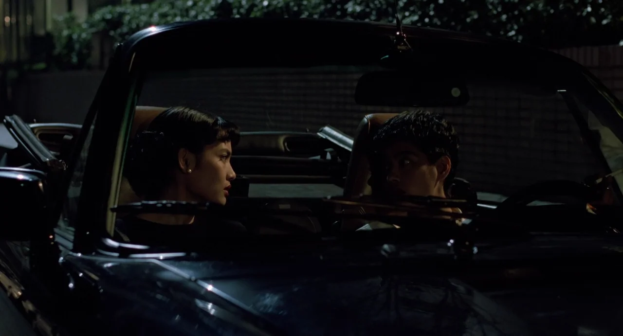 A Confucian Confusion (1994) movie still: medium — A dark convertible car is parked at night in a dimly lit outdoor space. A young woman and a man…