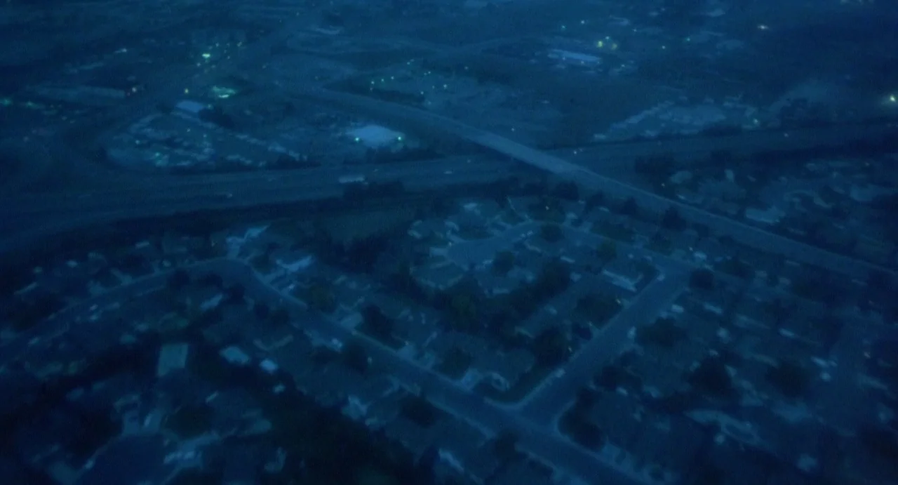 Explorers (1985) movie still: establishing — An aerial view shows a dense suburban housing development at night under blue light. A highway overpass…