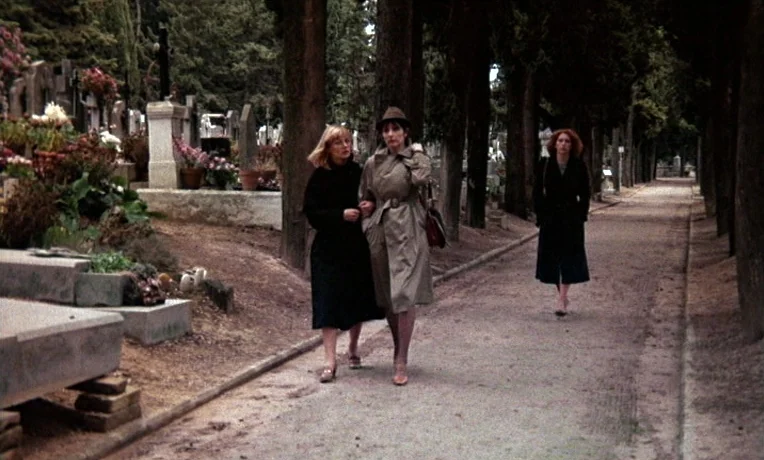 The Man Who Loved Women (1977) movie still: fullbody — A gravel path leads through an old cemetery flanked by dense, dark cypress trees. Two women…