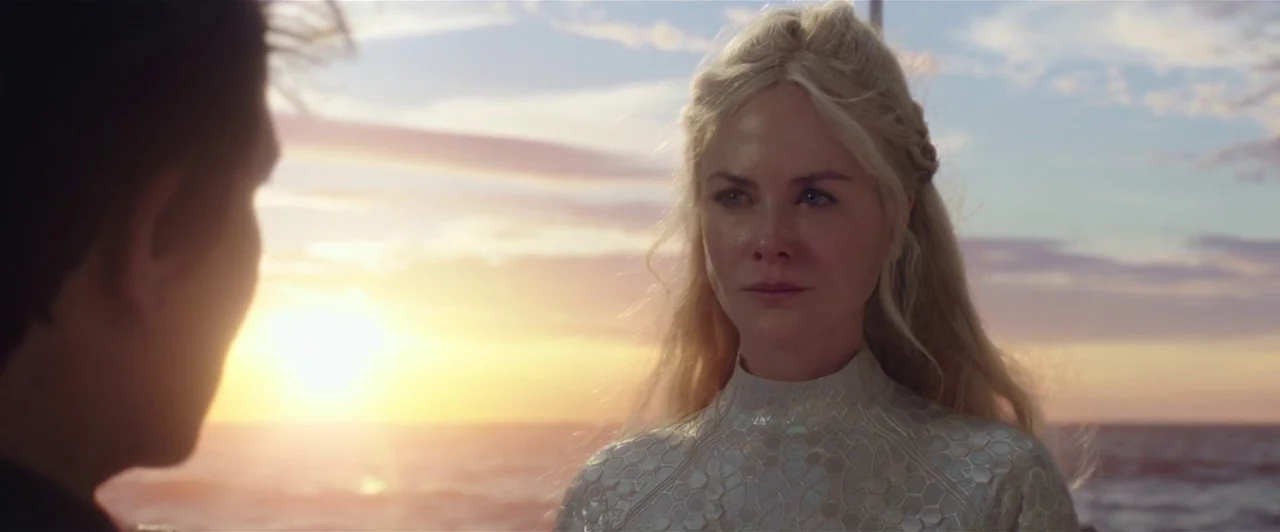 Aquaman (2018) movie still: medium — A woman with blonde hair stands near the ocean during the golden hour sunset. She looks toward a man whose…