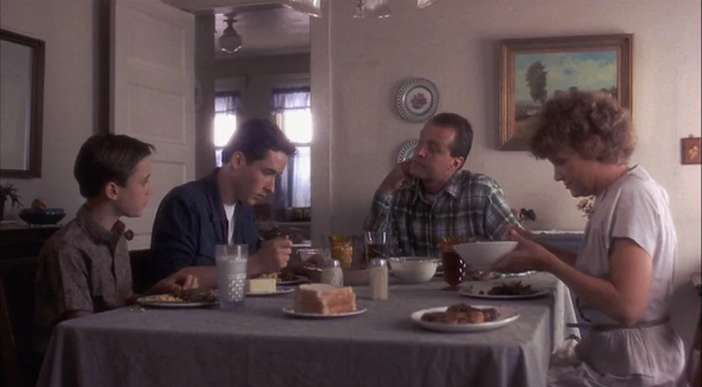 Stand By Me (1986) movie still: medium — A modest home interior with a dining table set for a family dinner. Four people sit around the table eating…