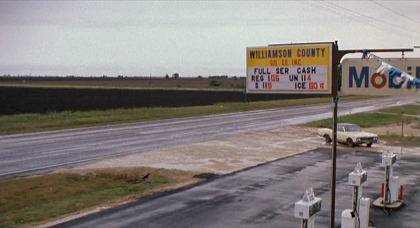 Blood Simple (1984) movie still: establishing — A rural gas station sits beside an empty asphalt highway under a grey sky. A yellow price sign…