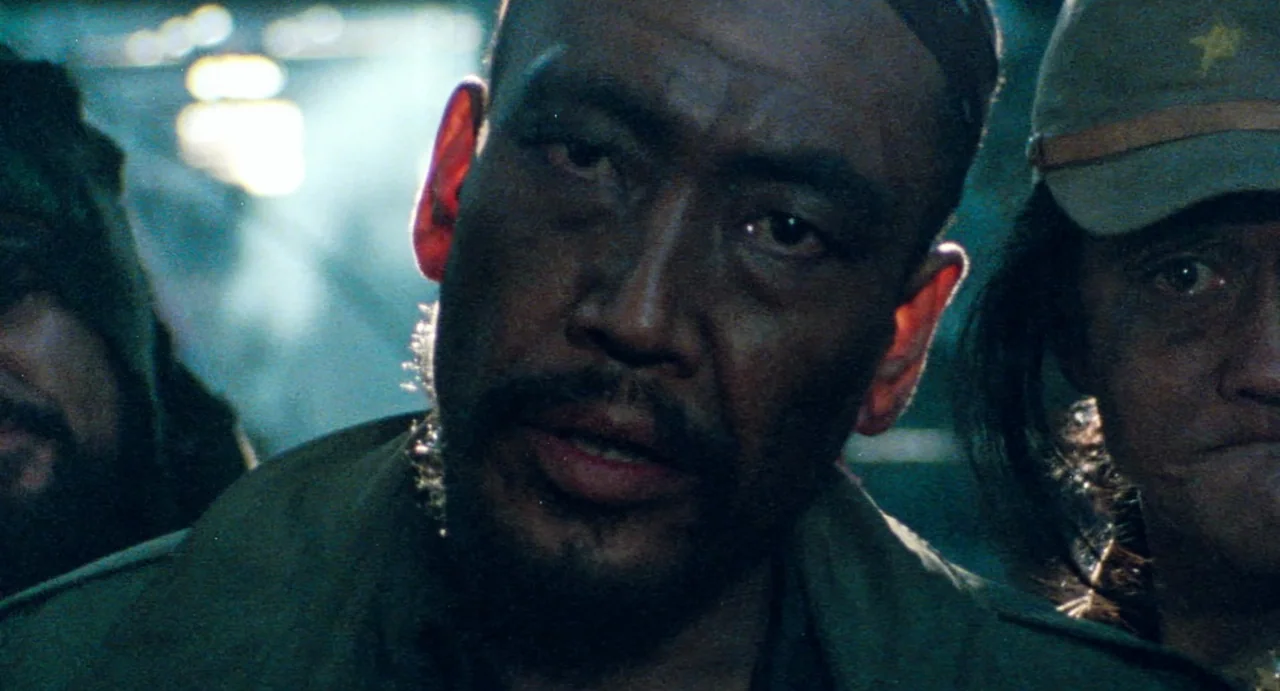Burst City (1982) movie still: closeup — A dense, dark jungle environment at night with low visibility and high moisture levels. A man looks toward…