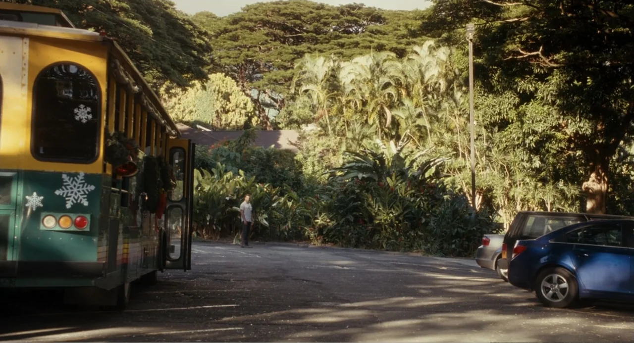 Aloha (2015) movie still: wide — A man stands in a parking lot adjacent to a tropical wooded area. A decorated holiday trolley bus is parked on the…