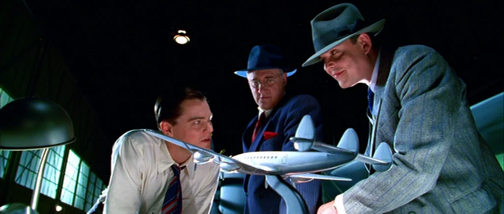 The Aviator (2004) movie still: medium — A dimly lit industrial interior space with large windows in the shadowed background. Three men in…