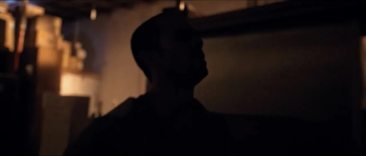 Gunman (2025) movie still: closeup — A dark indoor storage space is filled with stacked cardboard boxes in the background. A man stands in the…