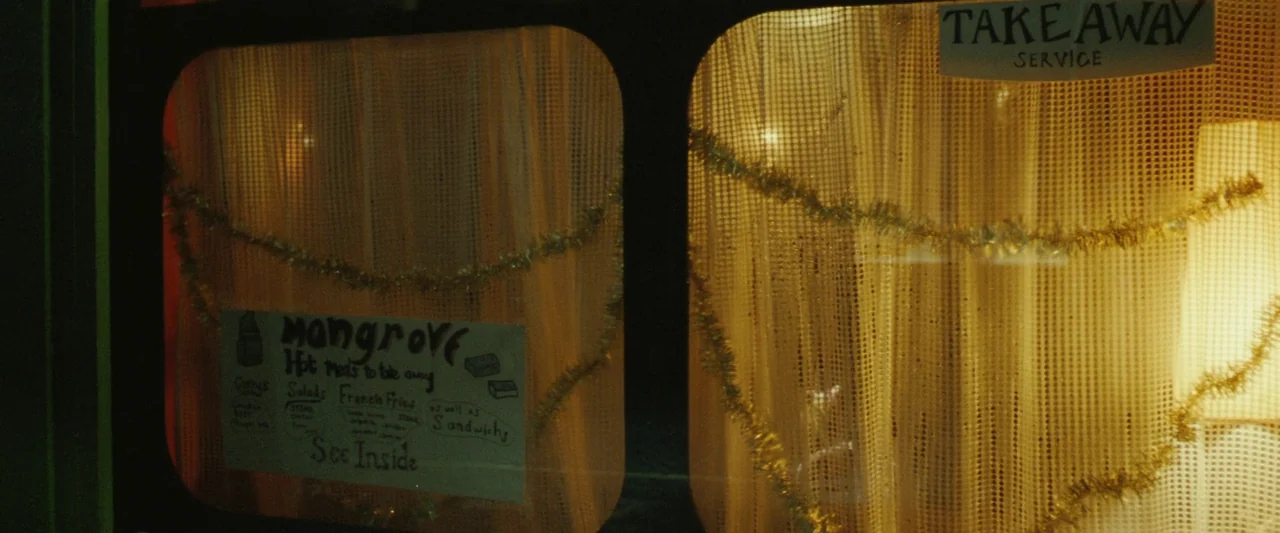 Mangrove (2020) movie still: wide — A restaurant window at night features gold tinsel and a handwritten takeaway menu sign. Warm yellow curtains…