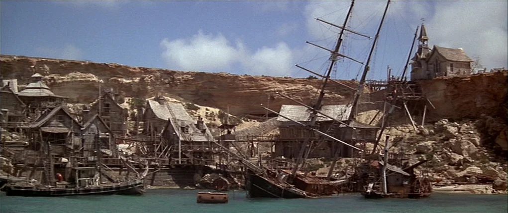 Popeye (1980) movie still: establishing — A rugged coastal settlement is nestled against a large, porous limestone cliff wall. Wooden houses and…