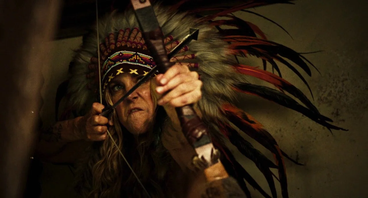 3 From Hell (2019) movie still: closeup — A woman in a dimly lit, nondescript interior space wears a large feathered headdress. She draws back a…