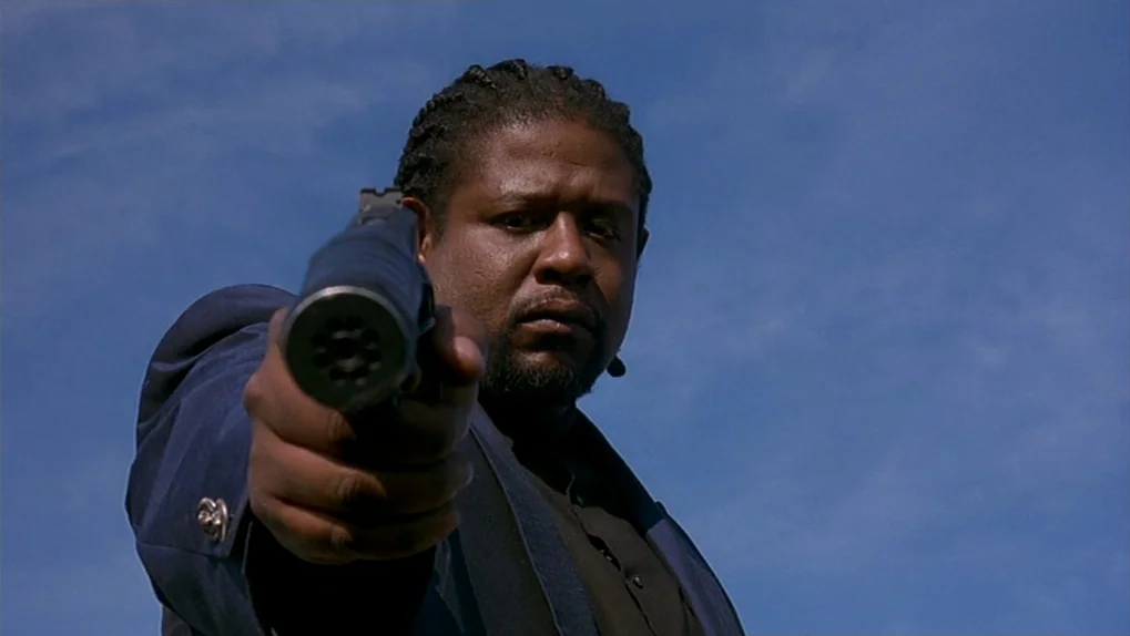 Ghost Dog: The Way of the Samurai (1999) movie still: closeup — A man stands outdoors under a clear blue sky. He aims a handgun directly toward the…