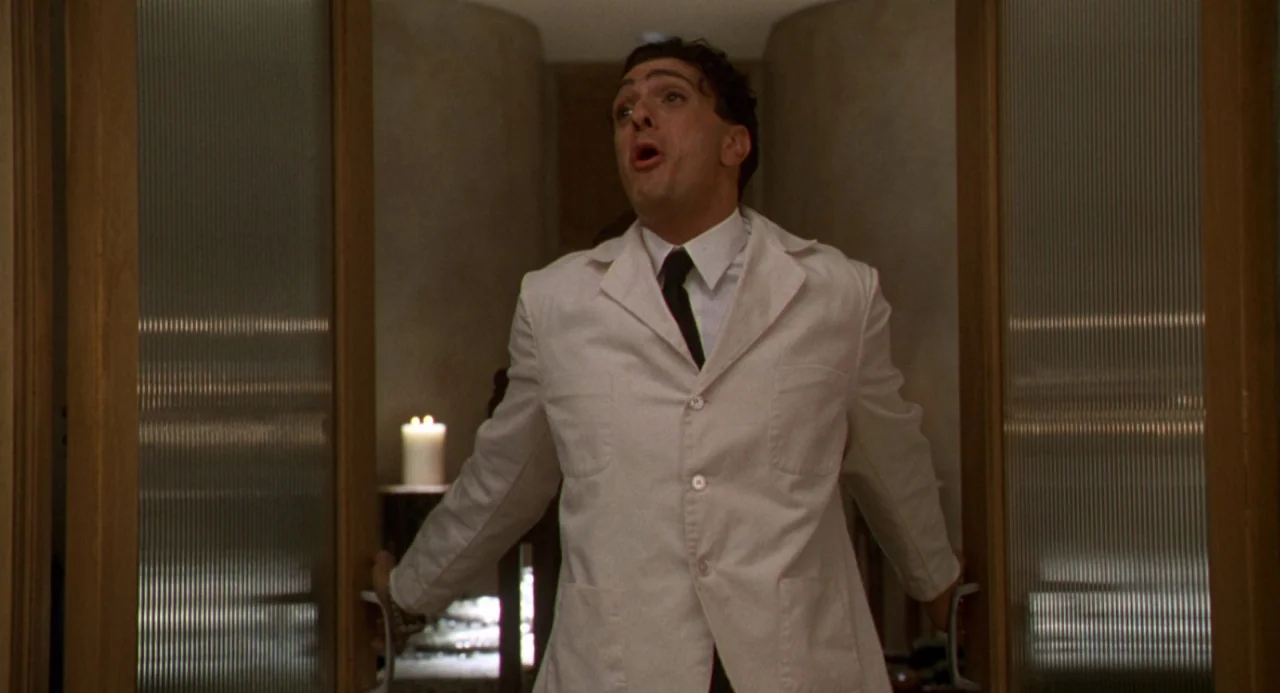 The Birdcage (1996) movie still: medium — A man in a white lab coat pulls open double sliding glass doors. A single lit candle stands on a small…